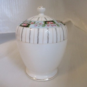 May include: White porcelain sugar bowl with a lid. The lid has a floral design with pink and green flowers on a blue background. The bowl has a gold trim and a black and white striped pattern.