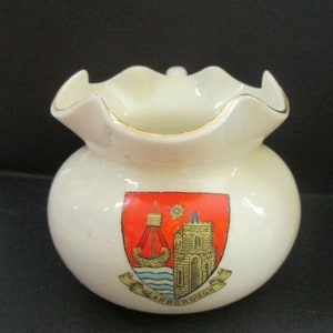 May include: A white ceramic creamer with a ruffled rim and a gold trim. The creamer features a coat of arms with a red background, a ship, a building, and a star. The text "SCARBOROUGH" is written on a banner below the coat of arms.