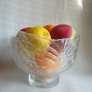 May include: A clear glass fruit bowl with a textured, frosted finish. The bowl has a wide, rounded base and a flared rim. It is filled with a variety of fruit, including lemons, oranges, and apples.