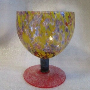 Pode incluir: Um copo de vidro colorido com um design salpicado de amarelo, vermelho, branco e azul. O copo tem um caule transparente e uma base vermelha.