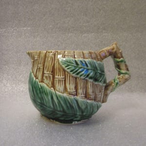 May include: A ceramic pitcher with a bamboo-style design. The body is tan with vertical lines, and the bottom is wrapped in green leaves. The handle is designed to look like a tree branch with green vines. A green leaf is on the side.
