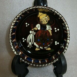 May include: A small, round ceramic dish with a dark brown glaze. The dish features a hand-painted design of a girl in a traditional dress holding a basket of flowers. The rim of the dish is decorated with a white and blue pattern.