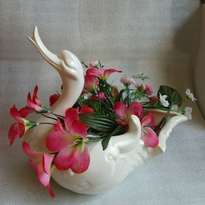 May include: A white ceramic swan planter with pink and white flowers. The swan's neck is curved upwards and the planter has a wide, open mouth.