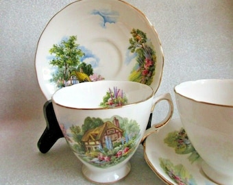 May include: A set of two vintage teacups and saucers with a cottagecore design. The cups and saucers feature a white background with a colorful illustration of a cottage surrounded by flowers and trees. The edges of the cups and saucers are trimmed with a gold band.