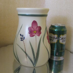 Puede incluir: Jarrón de cerámica color crema con flores rosas pintadas a mano y hojas verdes. El jarrón tiene una banda verde alrededor de la parte superior. Una lata verde de Canada Dry ginger ale está al lado del jarrón.