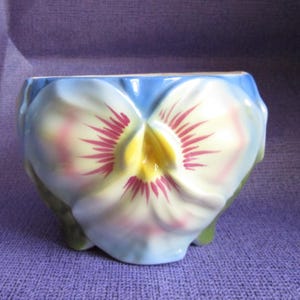 May include: A ceramic pansy flower-shaped planter. It has a blue rim, white petals with pink accents, a yellow centre, and green leaves. The design is hand-painted, a charming decorative item.