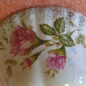 Könnte beinhalten: Nahaufnahme einer weißen Porzellantasse mit goldfarbenem Rand und einem Blumendesign mit rosa Rosen und grünen Blättern.