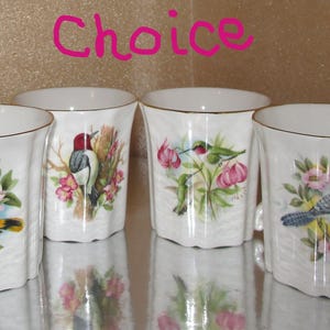 May include: Four white porcelain mugs with gold-trimmed rims and a textured, wavy surface. Each mug features a different colourful bird and floral illustration. The word Choice is written in pink text above the mugs in the centre of the image.