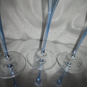 Puede incluir: Primer plano de varias copas de vino transparentes con un tinte azul claro. Los tallos son largos y delgados, y las bases son redondas. Las copas est&aacute;n dispuestas sobre una superficie reflectante.