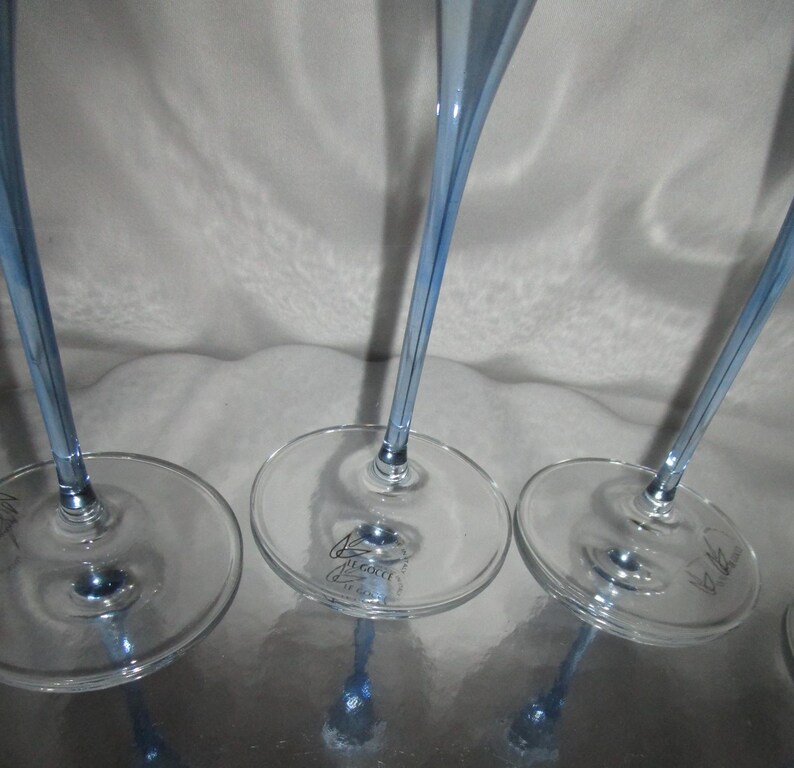 Puede incluir: Primer plano de varias copas de vino transparentes con un tinte azul claro. Los tallos son largos y delgados, y las bases son redondas. Las copas est&aacute;n dispuestas sobre una superficie reflectante.