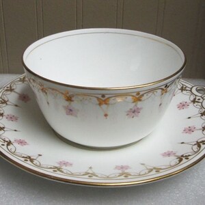 May include: A white porcelain teacup and saucer set with a delicate floral pattern in pink and gold. The cup and saucer have a scalloped edge and a gold trim.