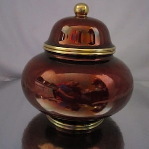 Könnte beinhalten: Ein dekoratives, rundes Gefäß mit Deckel. Das Gefäß hat eine tiefrote, braune Farbe mit glänzendem Finish und goldfarbenen Zierleisten. Der Deckel ist mit einem kleinen, goldfarbenen Knauf versehen. Das Gefäß steht auf einer reflektierenden Oberfläche.