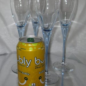 Puede incluir: Una lata amarilla de agua con gas Apple Bubly con cuatro copas de champ&aacute;n. Las copas tienen cuencos transparentes y tallos azul claro. La lata es de 355 ml.