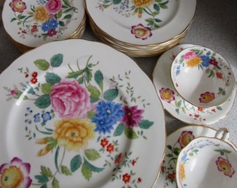 Puede incluir: Un juego de vajilla de cerámica blanca con un colorido estampado floral de flores rosas, amarillas y azules y hojas verdes. El conjunto incluye varios platos de distintos tamaños y dos tazas de té con sus platos, todos con borde dorado.