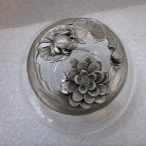 Op de afbeelding: Een heldere glazen kom met een decoratieve metalen rand. De rand heeft een kikker op een waterlelie, een bloem en een kleine vis. Het metaal heeft een zilveren afwerking. De kom is rond en heeft een brede opening.