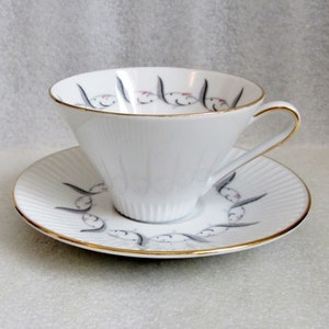 Op de afbeelding: Een wit theekopje en schotel met een gouden rand en een gestileerd vogelontwerp. De vogels zijn grijs en hebben roze accenten.