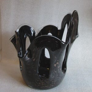 May include: A black glass vase with a wide, undulating rim and multiple large, oval-shaped cutouts along the sides. The glass features a dark, semi-translucent appearance with subtle, lighter-colored flecks embedded throughout the material.