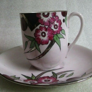 Puede incluir: Un juego de taza y platillo de té rosa con un diseño floral. La taza tiene un borde plateado y un asa. El platillo tiene un borde plateado y un diseño floral.