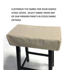 May include: A beige fabric saddle stool cover with black wooden legs. Customize the fabric for your saddle stool cover. Select fabric from any of our premier prints in stock fabric options.