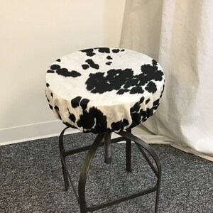 Pu&ograve; includere: Uno sgabello in pelle di mucca bianco e nero con base in metallo. Lo sgabello &egrave; su un tappeto grigio.