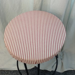 Puede incluir: Una funda de taburete redonda de tela a rayas rojas y blancas. La funda es redonda y se ajusta a un taburete de metal negro.