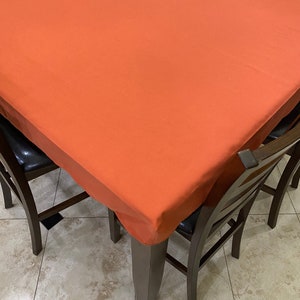 May include: A square dining table with a bright orange tablecloth and four chairs. The table and chairs are made of dark wood.