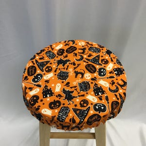 Könnte beinhalten: Ein runder Hockerbezug aus orangefarbenem Stoff mit einem schwarz-weißen Halloween-Muster. Der Druck zeigt Illustrationen von Hexen, Kürbissen, Geistern, Totenköpfen, Spukhäusern, Katzen sowie den Text Happy Halloween und Fright Night.