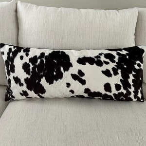 May include: A long, rectangular throw pillow with a black and white cowhide pattern. The pillow is sitting on a white couch.