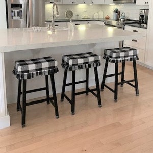 May include: Three black and white checkered pattern bar stools with black legs are positioned in front of a white kitchen island. The stools have a ruffled trim around the seat. The kitchen has white cabinets and stainless steel appliances.