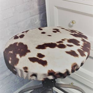 May include: A brown and white cowhide patterned chair seat cover. The cover is made of a soft, plush fabric and has a gathered edge.