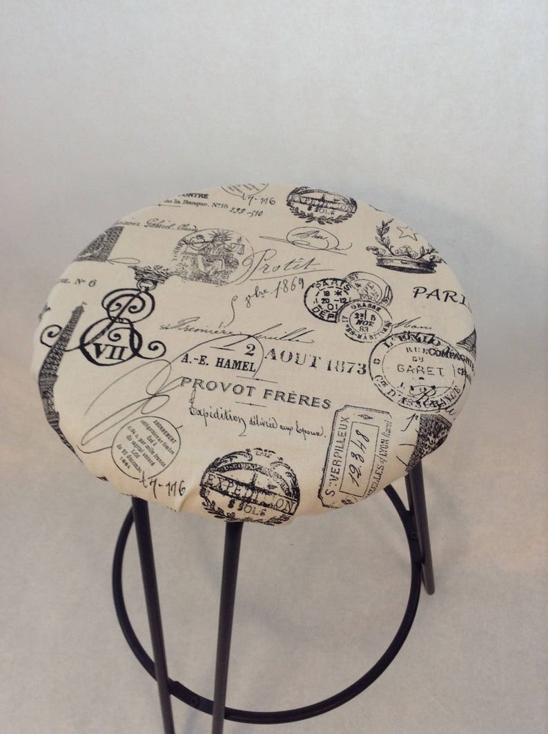 Pu&ograve; includere: Uno sgabello beige con struttura in metallo nero. La seduta &egrave; rivestita in tessuto con un design parigino vintage con timbri e scritte nere.