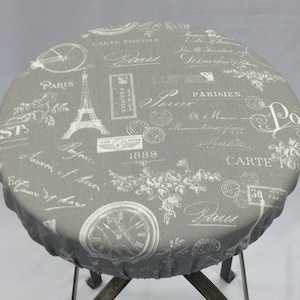 May include: Grey round fabric table cover with a white Parisian-themed design. The design features the Eiffel Tower, bicycles, clocks, and the words "Carte Postale" and "Paris".