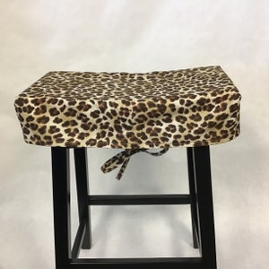May include: A brown and beige leopard print stool cover with a tie closure. The cover is made of fabric and fits over a black stool.