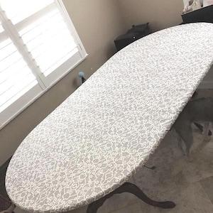 May include: An oval-shaped table cover featuring a repeating white and light gray damask-style pattern. The fabric is fitted tightly over the surface of an oval table with dark wooden legs, creating a smooth and uniform appearance across the top.