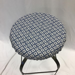 Op de afbeelding: Een blauwe en witte stoffen stoelhoes met een gerimpelde rand. De hoes is op een zwarte metalen kruk met een ronde zitting.