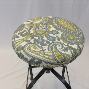 May include: A round, white fabric stool cover with a paisley pattern in shades of grey, yellow, and blue. The cover is fitted over a black metal stool.