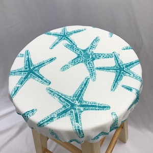 Puede incluir: Una funda redonda para taburete con base de tela blanca y un patrón repetitivo de estrellas de mar color verde azulado. La funda tiene un borde elástico que se ajusta al asiento circular de un taburete de madera clara de cuatro patas.