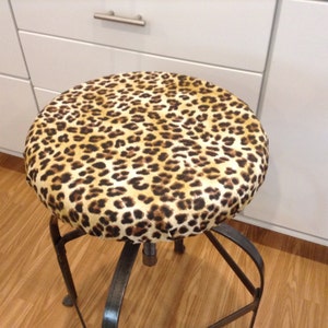 May include: A leopard print stool cover with a brown, black and beige pattern. The cover is made of fabric and fits over a metal stool.
