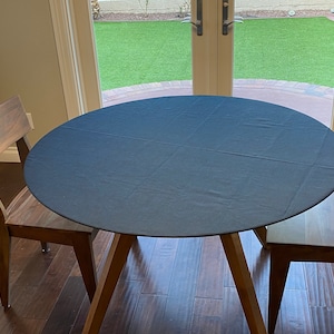 May include: A round, dark charcoal gray fabric tablecloth covering a circular wooden table. The tablecloth features visible seam lines forming a cross pattern across the surface, and the table is accompanied by two matching wooden chairs.