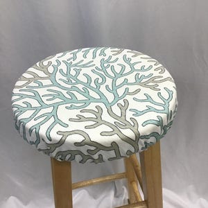 May include: A round wooden stool featuring a padded seat cover with a white background and a repeating pattern of branching coral in shades of light blue and grey. The stool has four straight wooden legs and a simple, functional design.