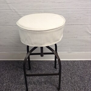 May include: A white fabric stool cover with a black metal base. The cover is round and has a simple design. The base has a swivel function.