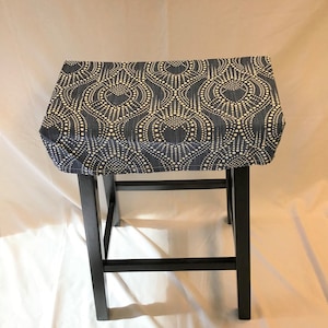 May include: A black wooden stool with a rectangular seat covered in a navy blue fabric. The fabric features a repeating pattern of cream-colored circles and dots. The stool has four black legs and a footrest.