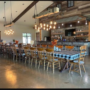 May include: A large wooden dining table with a black and white checkered tablecloth is set with chairs in a rustic dining room. The room has a bar area with a wooden countertop and shelves stocked with bottles. The ceiling has exposed wooden beams and hanging light fixtures.