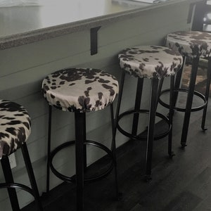 May include: Four black metal bar stools with cowhide patterned seat covers. The stools have a simple design with a round seat and a single ring footrest.