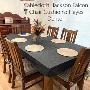 May include: A dining table set with a dark grey tablecloth and woven placemats. Wooden chairs with striped cushions are arranged around the table. A floral arrangement in a vase sits in the centre. Text on the image reads: Tablecloth: Jackson Falcon, Chair Cushions: Hayes Denton.