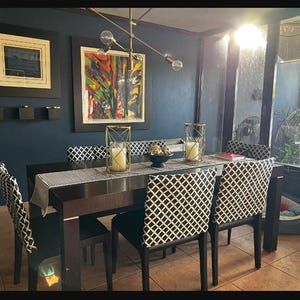 May include: A dark blue dining room with a black and white patterned dining table and chairs. The table has a black runner and a gold candle holder. There is a large abstract painting on the wall behind the table.