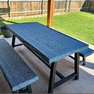 May include: A rectangular outdoor dining table and bench set. The table and bench have black legs and are covered with a textured, blue fabric. The set is on a concrete patio with a green lawn and wooden fence in the background.