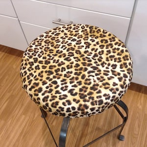 May include: A leopard print stool cover with a brown and tan pattern. The cover is made of fabric and fits over a round stool.