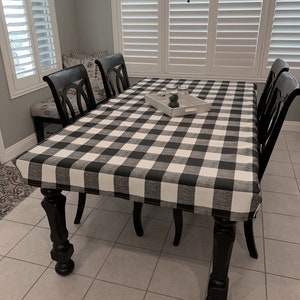 May include: Black and white checkered tablecloth with a white tray and two small jars on top of it. The tablecloth is on a black wooden table with four black chairs.