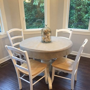 May include: A white round table with a light gray tablecloth and four white chairs with natural wood seats. A birdcage with white and gold decorative balls sits in the center of the table.